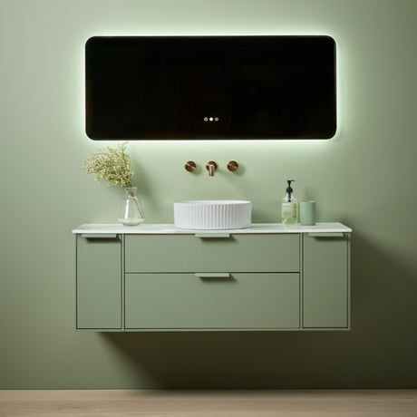 Modern sage green wall-mounted bathroom vanity unit with white countertop, integrated white ceramic basin, copper wall-mounted faucet, illuminated LED mirror with rounded corners, and decorative items including glass vase with baby's breath flowers and soap dispenser