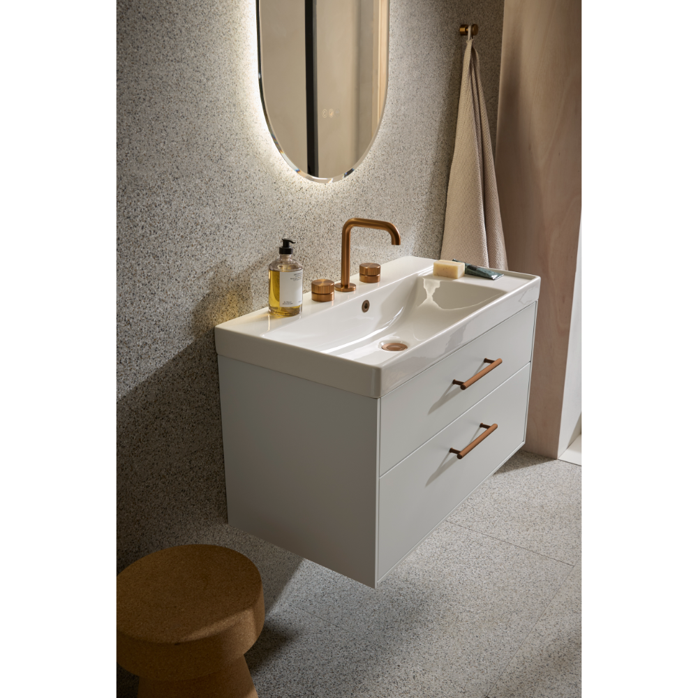 Modern white wall-mounted bathroom vanity unit with dual ceramic sink, brass faucet, two storage drawers with copper handles, illuminated oval mirror, terrazzo flooring, and beige towel rail
