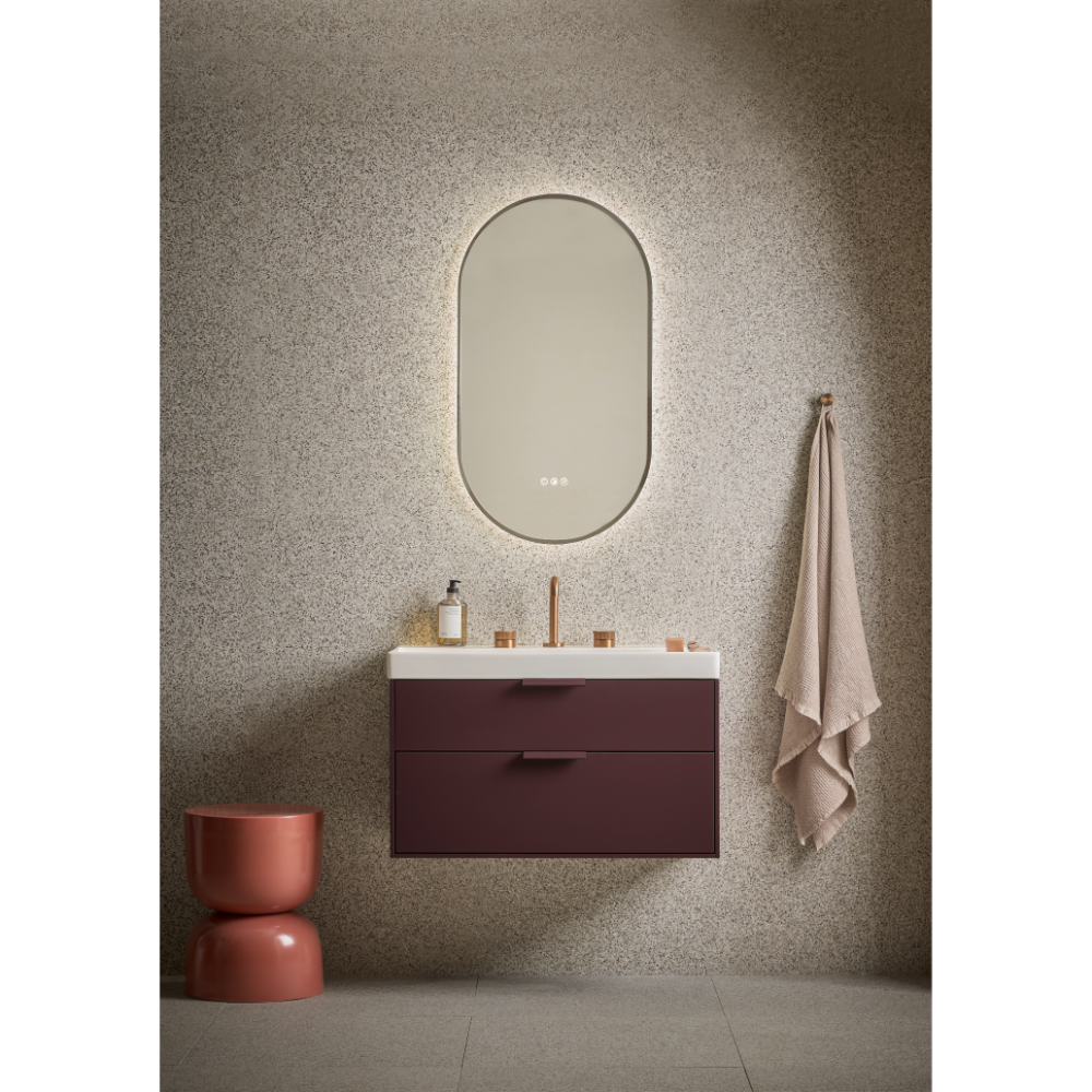 Modern bathroom vanity setup featuring a burgundy Roper Rhodes Faber wall-mounted vanity unit with white countertop and integrated sink, paired with an illuminated oval mirror with soft gold lighting, terracotta ceramic accessories, and neutral beige towel against textured taupe wall
