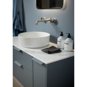 Modern bathroom vanity unit with white marble countertop, dark grey cabinetry, round white ceramic vessel sink, brass wall-mounted faucet, two pump dispensers, dark grey towel and mirror above