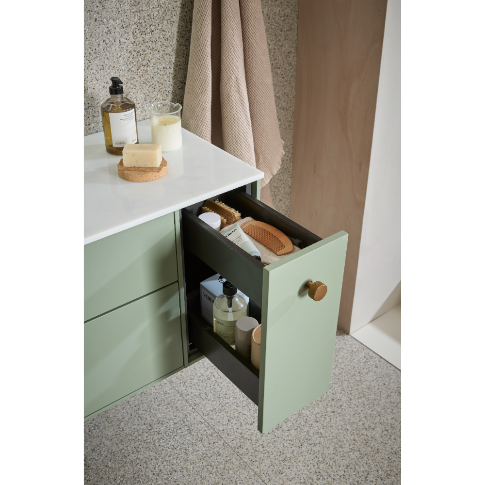 Sage green and black vanity unit with white countertop featuring open drawer displaying bathroom toiletries, soap, brushes and bottles, wooden handle, terrazzo flooring and neutral fabric curtains in background