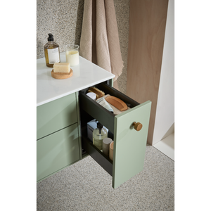 Sage green and black vanity unit with white countertop featuring open drawer displaying bathroom toiletries, soap, brushes and bottles, wooden handle, terrazzo flooring and neutral fabric curtains in background
