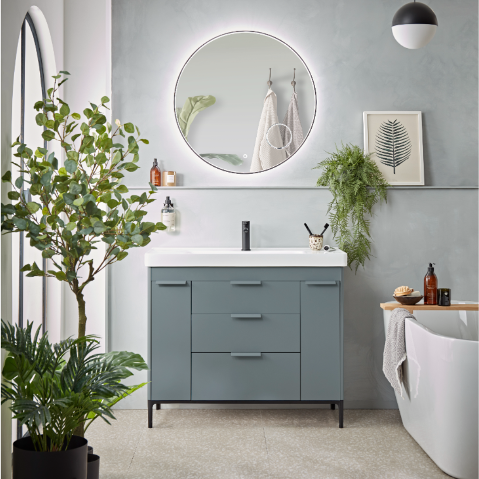 Modern bathroom interior featuring a matte carbon Fairmont vanity unit with white countertop and black metal legs, round illuminated mirror, matte agave green freestanding bathtub, grey walls, potted plants, pendant lighting, and botanical wall art
