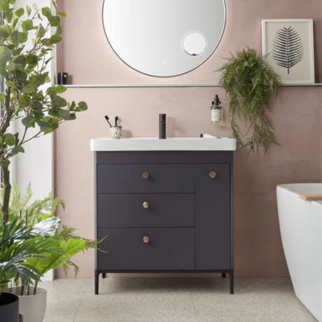 Modern bathroom vanity unit with matte carbon finish featuring white countertop, round mirror with chrome frame, wooden handles, integrated sink and black faucet, surrounded by potted green plants and ferns against soft pink walls with beige flooring