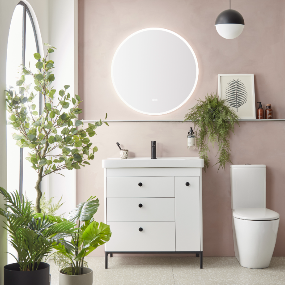 Modern bathroom vanity unit in gloss white with matte carbon handles, paired with a circular illuminated mirror, matching toilet, and decorative plants against a mauve wall