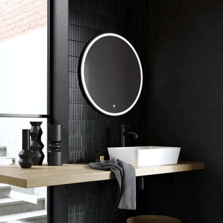 Modern black bathroom with round illuminated LED mirror above white vessel sink on light wood countertop, black wall tiles, and matte black fixtures