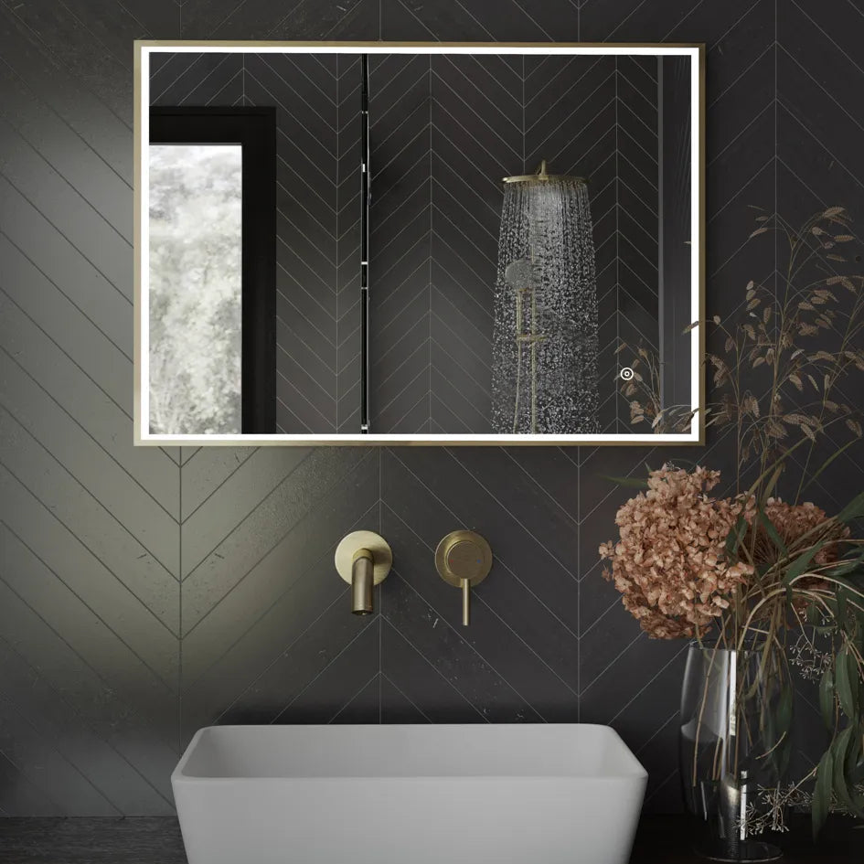 Modern bathroom vanity featuring Aqualla Fuse Frame LED mirror with brushed brass frame and black herringbone tile wall, white vessel sink, gold wall-mounted faucet, rainfall showerhead, and dried flower arrangement