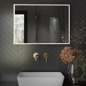 Modern bathroom vanity featuring Aqualla Fuse Frame LED mirror with brushed brass frame and black herringbone tile wall, white vessel sink, gold wall-mounted faucet, rainfall showerhead, and dried flower arrangement