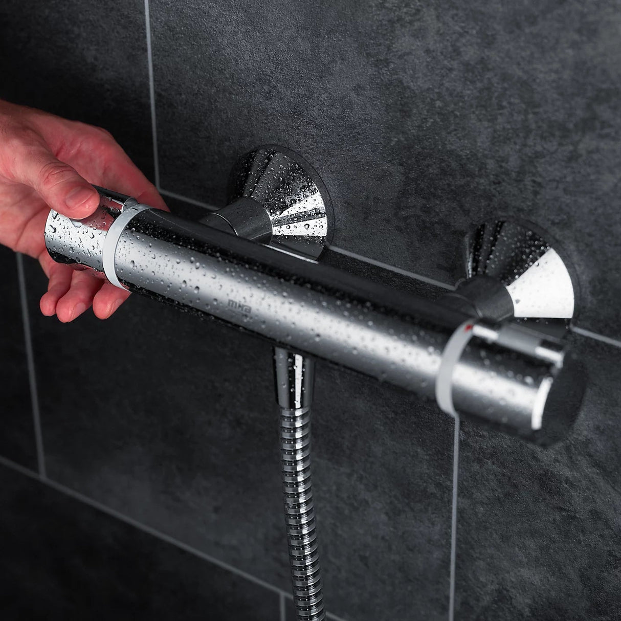 Mira Relate exposed bar mixer shower in polished chrome mounted on dark grey tile wall with water droplets on showerhead and hand adjusting controls
