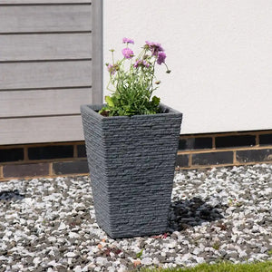 Strata Slate Tall Garden Planter Pot in ash grey with textured brick-pattern finish containing pink flowering plants, positioned on white and grey gravel beside a modern house with grey cladding