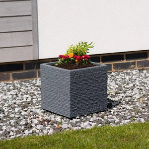 Strata Slate square garden planter pot in ash grey with textured brick-effect finish containing red, orange and pink flowering plants and green foliage, positioned on white gravel in a modern garden setting against a white wall