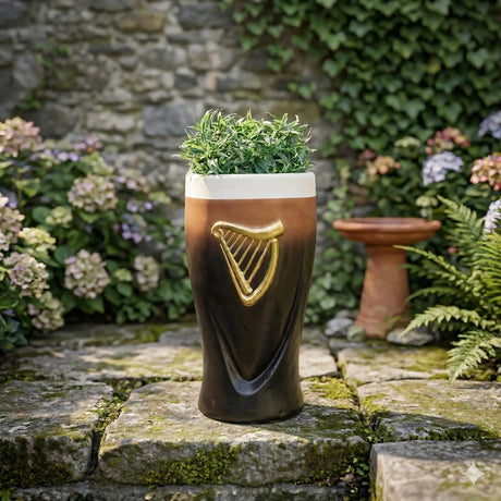 Black ceramic pint glass planter with gold harp emblem and cream foam head top, filled with green foliage, displayed on moss-covered stone in a garden setting surrounded by flowering plants and ivy