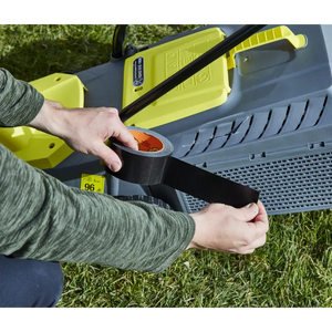 Person applying black Gorilla Tape to yellow and grey tool equipment outdoors on grass, showing the tape's adhesive strength and durability in practical use