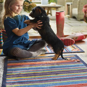 Young girl sitting on a colorful striped Hug Rug runner mat playing with a black dachshund dog in a sunny outdoor patio setting