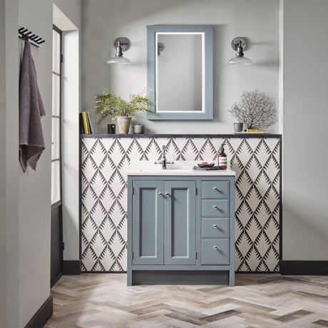 Modern bathroom vanity unit in matt slate grey with white ceramic sink, featuring dual chrome wall sconces and illuminated mirror with blue-grey frame, set against geometric patterned tile backsplash in grey and white, herringbone wood-look flooring, and styled with potted plants and bathroom accessories