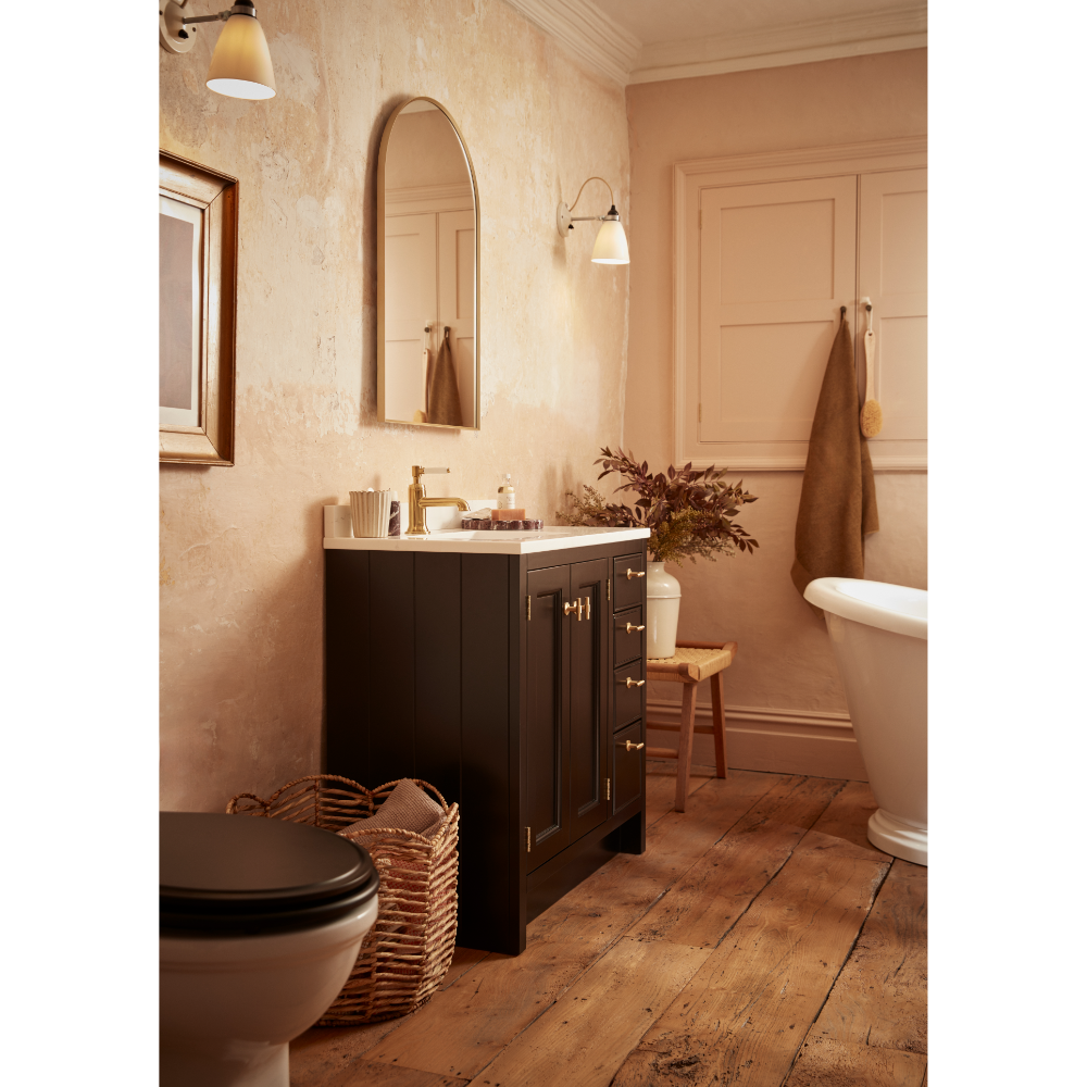 Matt Black Hampton vanity unit with white countertop in warm peach bathroom featuring wooden flooring, arched mirror, brass fixtures and traditional styling