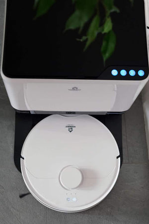 White Homebot Glider robotic vacuum cleaner with circular disc design and central button, shown from top view displaying blue LED indicators and docking station above, with green plant foliage visible in background