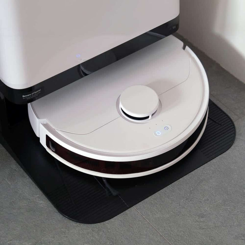 White and black robotic vacuum cleaner with circular disc design, dust collection bin on top, and control buttons, positioned on black mat against grey floor