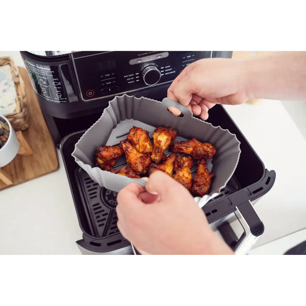Person holding open a black air fryer basket containing cooked golden brown chicken pieces lined with a gray silicone air fryer liner, displaying the product in use