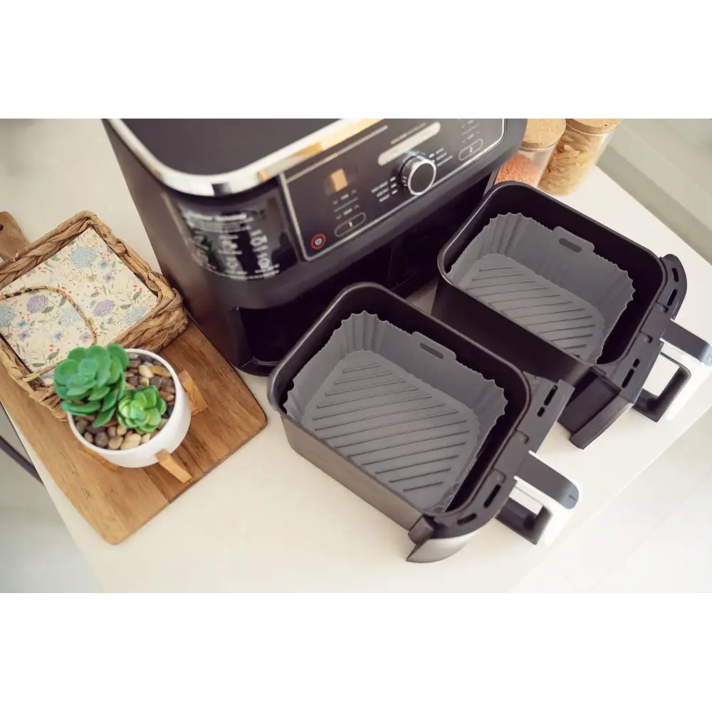 Two gray silicone air fryer liners displayed next to a black air fryer appliance, with a wooden cutting board holding decorative items including a succulent plant and woven basket in the background, on a white countertop