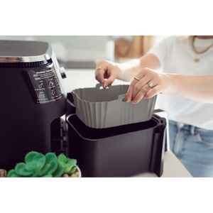Woman in white shirt placing gray silicone air fryer liner into black air fryer basket next to air fryer unit with succulent plant visible in foreground