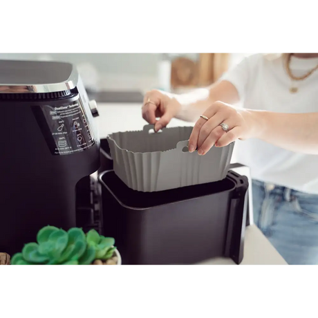 Woman in white shirt placing gray silicone air fryer liner into black air fryer basket next to air fryer unit with succulent plant visible in foreground