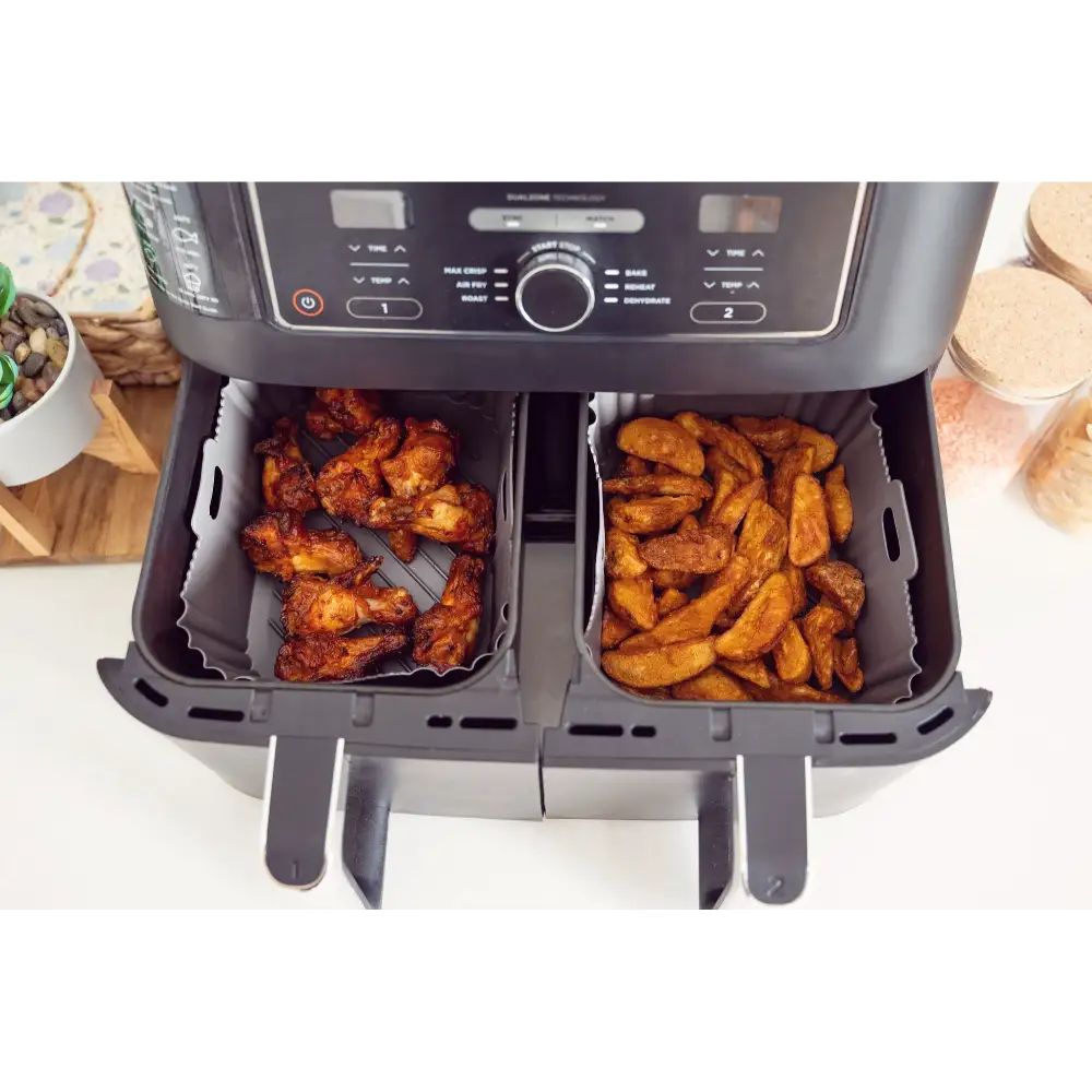 Dual basket air fryer with silicone liners containing golden-brown fried chicken wings on left basket and crispy potato wedges on right basket, viewed from above on white countertop with food prep items surrounding the unit