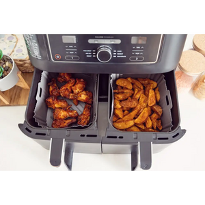 Dual basket air fryer with silicone liners containing golden-brown fried chicken wings on left basket and crispy potato wedges on right basket, viewed from above on white countertop with food prep items surrounding the unit
