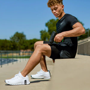 Young male athlete in black Nike sports shirt and shorts wearing white sneakers sits poolside using a black Hyperice Hypervolt Go 3 massage gun on his arm for muscle recovery