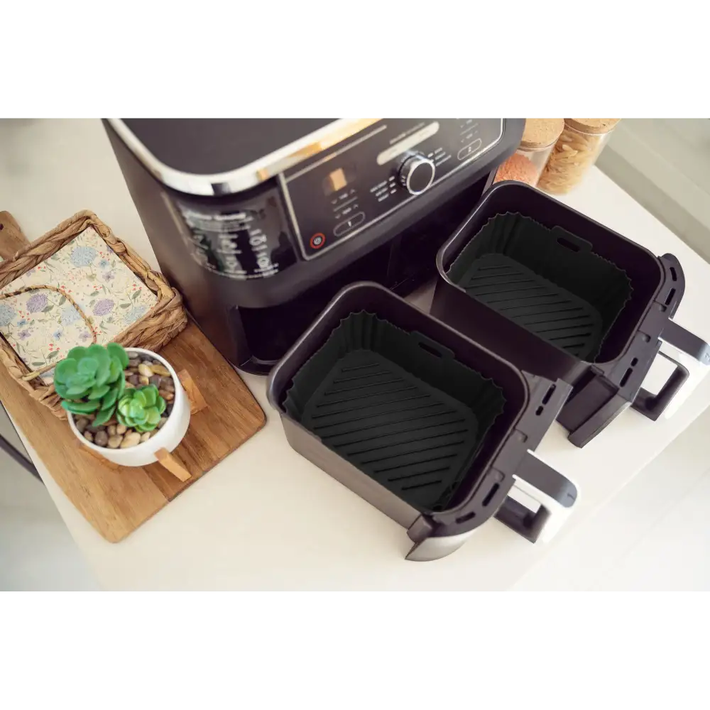 Two black silicone air fryer liners with ridged bottoms displayed next to a dual-basket air fryer, alongside a wooden cutting board with succulent plant and decorative items on a white countertop