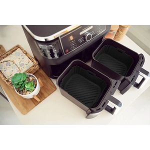 Two black silicone air fryer liners with ridged bottoms displayed next to a dual-basket air fryer, alongside a wooden cutting board with succulent plant and decorative items on a white countertop