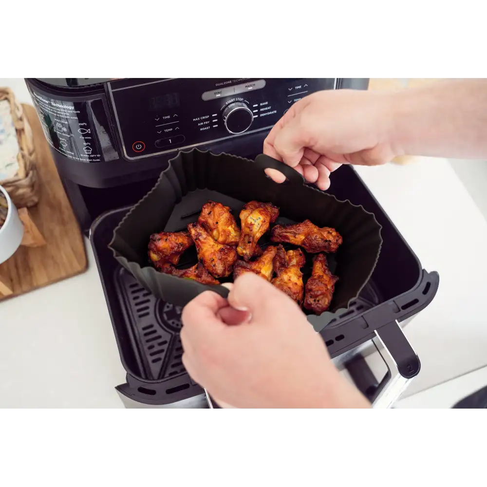 Hands removing a black silicone air fryer liner filled with cooked golden-brown chicken pieces from a black air fryer appliance on a white kitchen counter