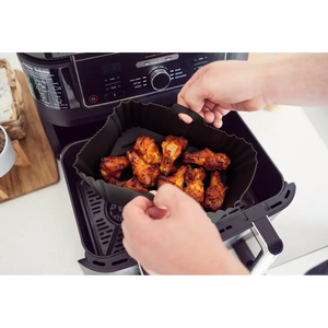 Hands removing a black silicone air fryer liner filled with cooked golden-brown chicken pieces from a black air fryer appliance on a white kitchen counter