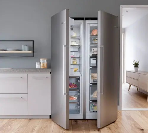 A tall stainless steel side-by-side refrigerator with both doors open, showing the interior shelves and freezer drawers stocked with food, set in a modern kitchen with light wood floors and white cabinetry.