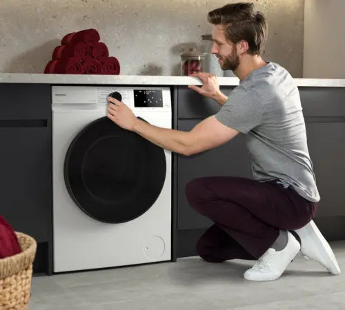 A man in a gray t-shirt and burgundy pants kneels down next to a white Siemens washing machine, touching its control panel in a modern laundry room setting.