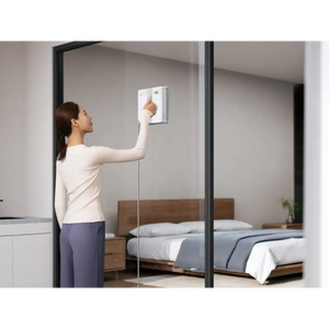 Woman in cream long-sleeve top and navy pants operating Ecovacs Winbot Mini white robot window vacuum on bedroom mirror, with wooden bed frame and gray bedding visible in reflection