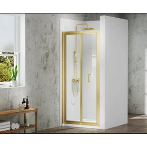 Merlyn MBOX brushed brass bifold shower door with clear glass panels in modern bathroom setting featuring concrete walls, wooden shelf with toiletries, and potted plant by window