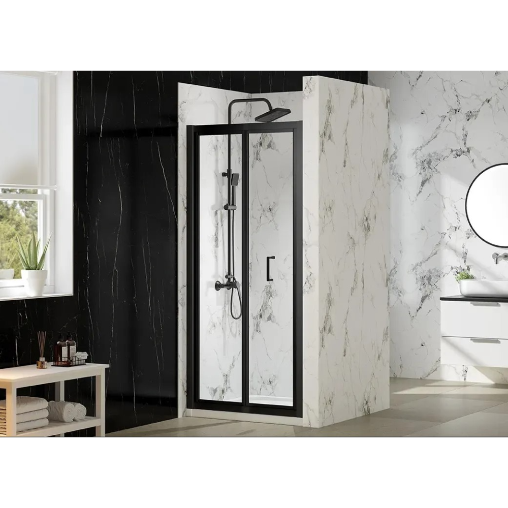 Modern bathroom featuring a Merlyn MBOX bifold shower door with matt black frame and clear glass panels, installed in a marble-effect enclosure with white and grey veining, alongside black marble wall tiles, circular mirror, white sink unit and wooden shelf with toiletries