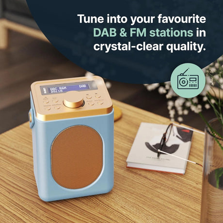 Majority Little Shelford portable DAB radio in duck egg blue and wood finish displaying BBC Radio on its LCD screen, sitting on a wooden table with flowers and a book, showcasing crystal-clear DAB and FM tuning capability