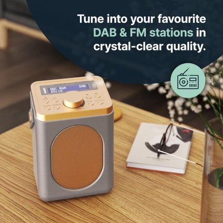 Majority Little Shelford portable DAB radio in grey and gold on wooden table with display showing BBC Radio station, featuring large tan speaker grille, control knob, and buttons for tuning DAB and FM stations in crystal-clear quality