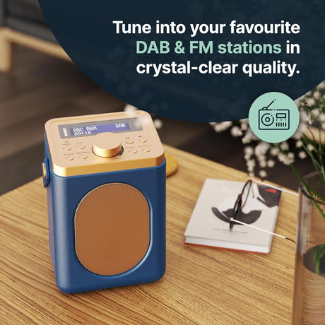 Majority Little Shelford portable DAB radio in blue and gold with cream speaker grille, displaying BBC Radio on LCD screen, sitting on wooden table with notebook and potted plant