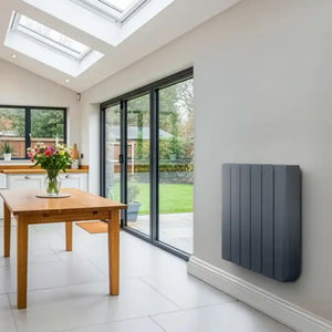 Grey wall-mounted oil filled radiator in modern kitchen extension with wooden dining table, white cabinetry, skylights and black framed glass doors overlooking garden