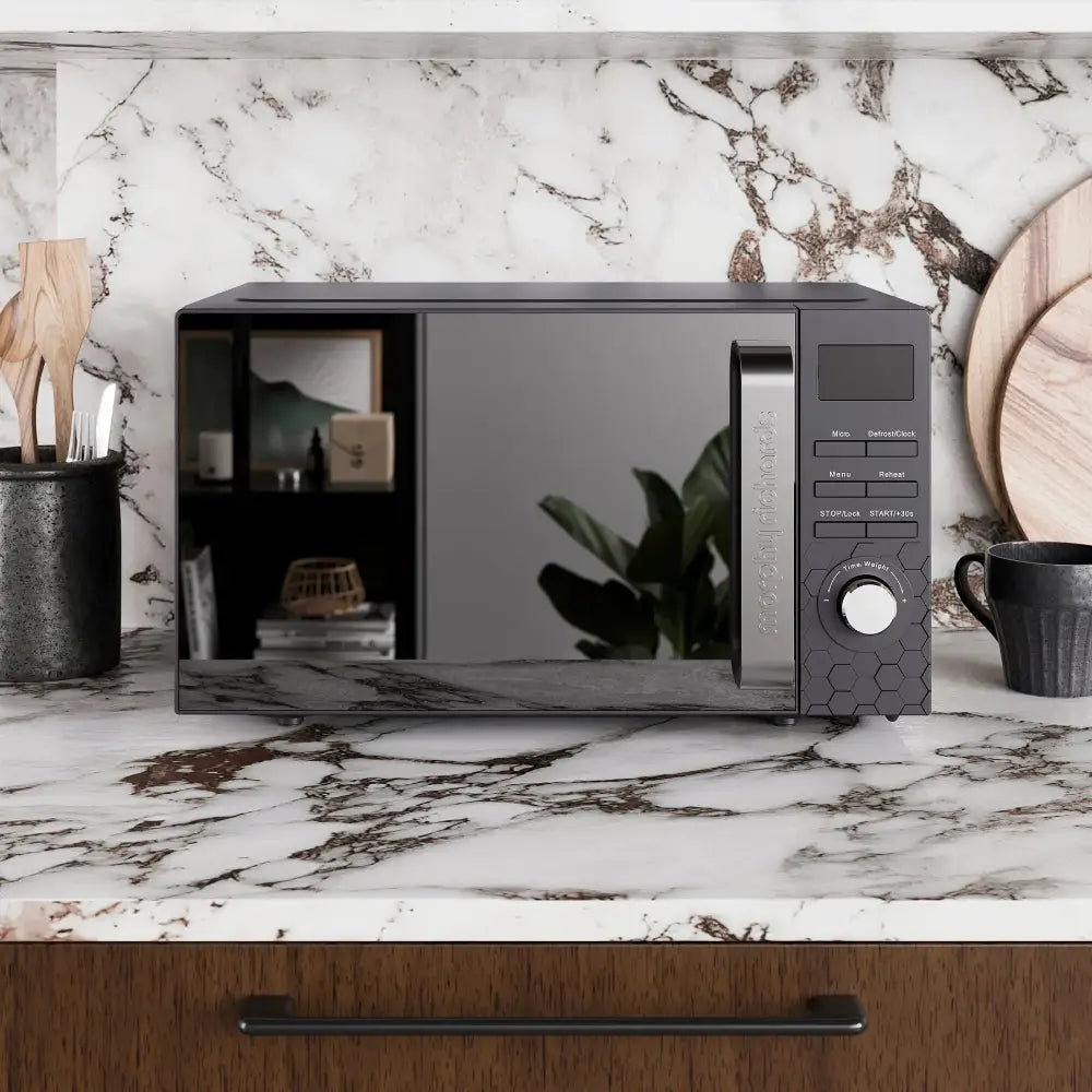 Black Morphy Richards Cassini digital microwave with 20 litre capacity positioned on a marble countertop with veining, surrounded by black ceramic containers and wooden accessories, featuring a digital display panel and control dial on the right side