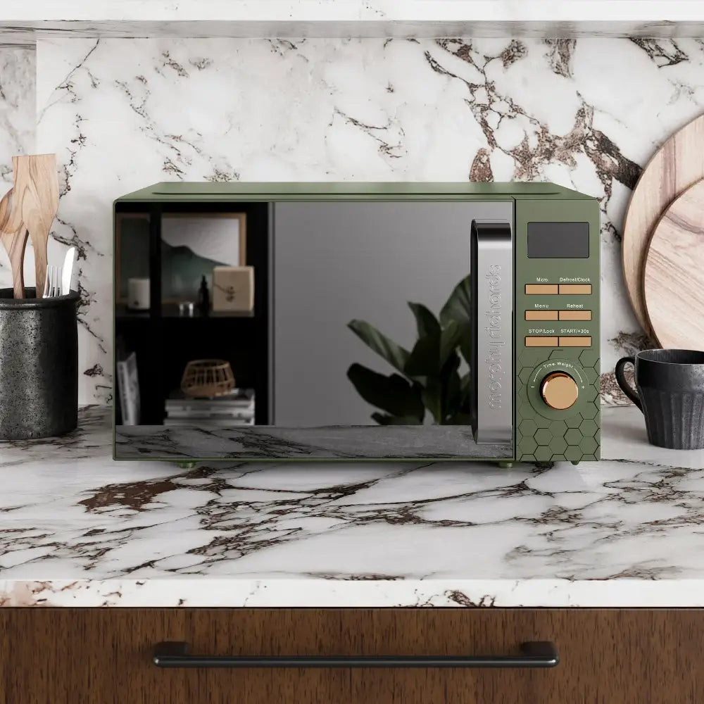 Morphy Richards Cassini 20 litre digital microwave in sage green and black with honeycomb detail, displayed on a marble countertop with black ceramic containers and wooden accessories in a modern kitchen setting