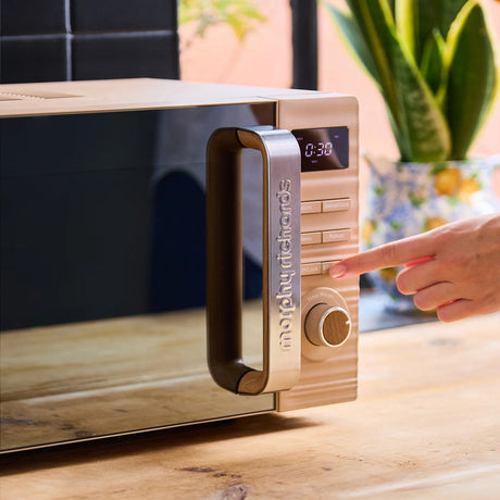 Morphy Richards Clay 20 Litre 800w microwave in cream and wood finish with digital display showing time, positioned on wooden kitchen counter with potted plant and person's hand operating the control dial
