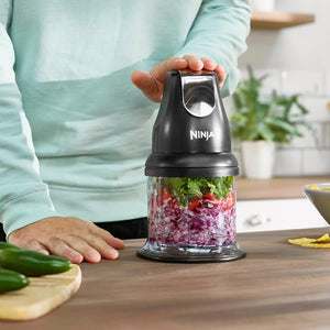 Person in light blue long-sleeved top holding black Ninja Professional Stackable Chopper 200W NJ1002UKBK with chopped red onions and fresh herbs visible in clear container, wooden kitchen counter with fresh vegetables and potted plant in background