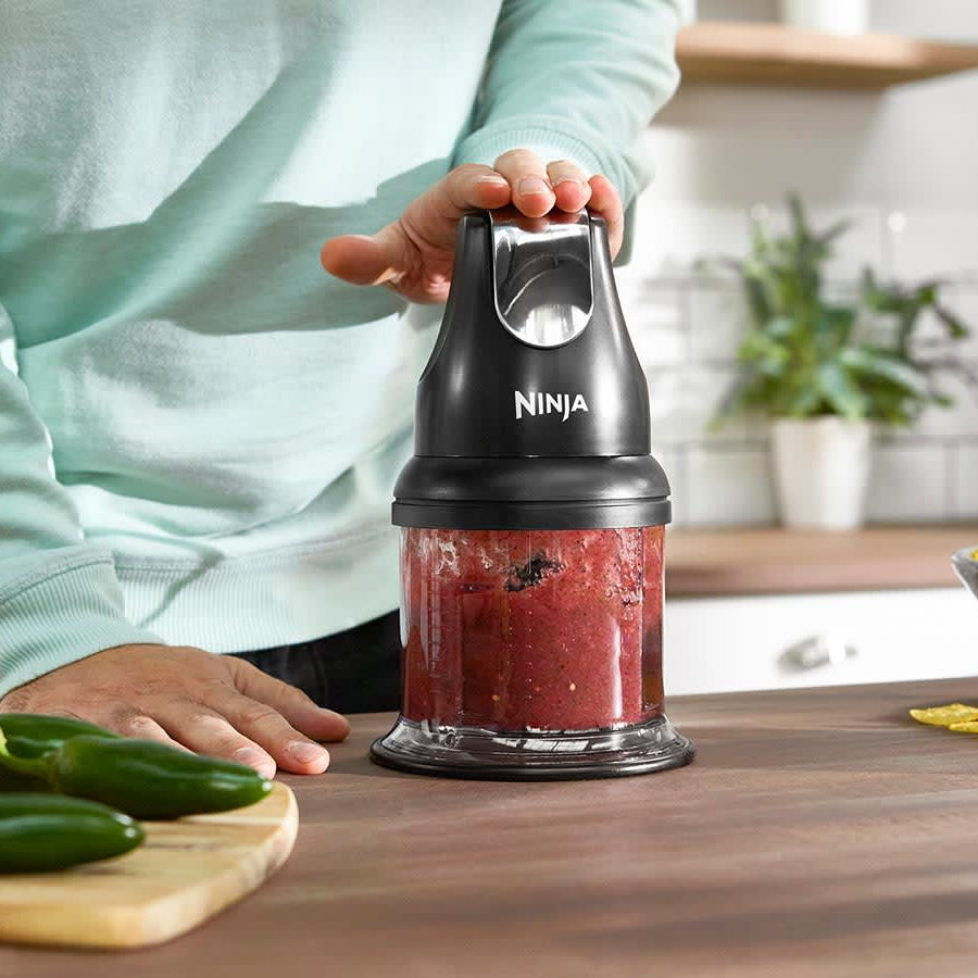 Person in mint green sweater holding black Ninja Professional Stackable Chopper with red chopped ingredients visible in the clear bowl, wooden kitchen counter with fresh cucumbers and potatoes nearby, white shelving and potted plants in background