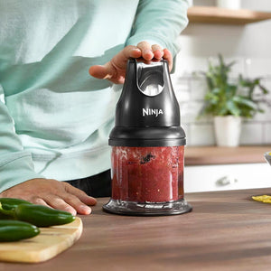 Person in mint green sweater holding black Ninja Professional Stackable Chopper with red chopped ingredients visible in the clear bowl, wooden kitchen counter with fresh cucumbers and potatoes nearby, white shelving and potted plants in background