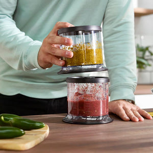 Person in mint green long-sleeved top holding Ninja Professional Stackable Chopper with two clear processing bowls stacked together, top bowl containing yellow chopped ingredients and bottom bowl containing red chopped ingredients, black plastic lids and base, wooden countertop with green peppers on cutting board visible in foreground