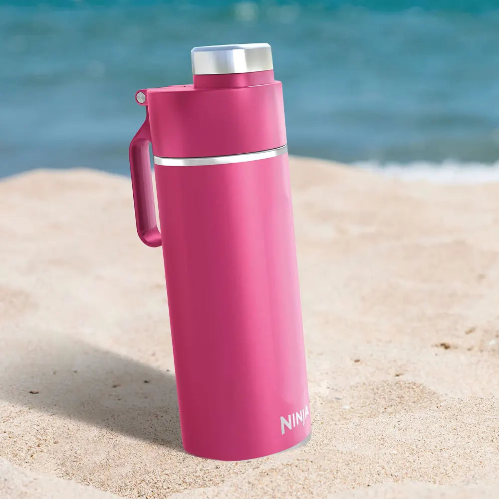 Ninja Thirsti 530ml insulated travel bottle in hot pink with silver trim and handle, positioned on sandy beach with ocean water in background